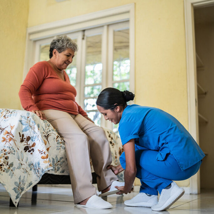 Mid Adult Nurse Helping Senior Woman Put On Slippers In Bedroom In Nursing Home